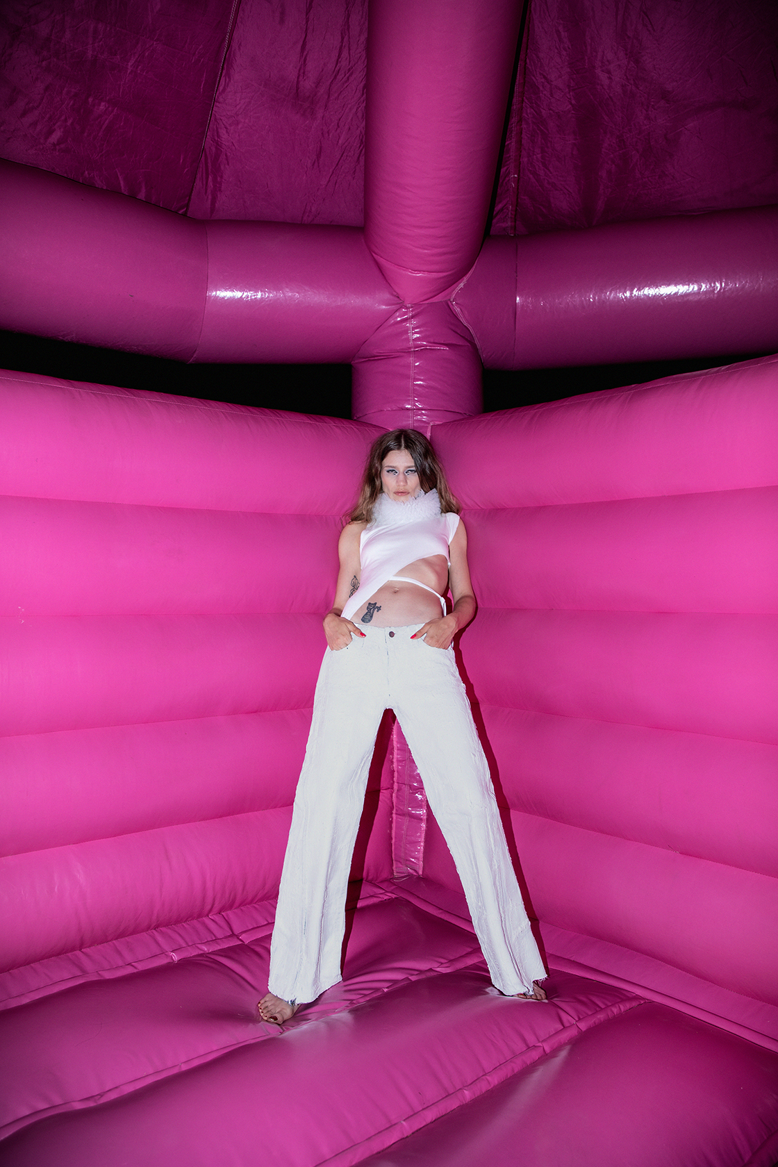 Model in white pants and top leaing against the wall of a bright pink bounce house glaring at the camera